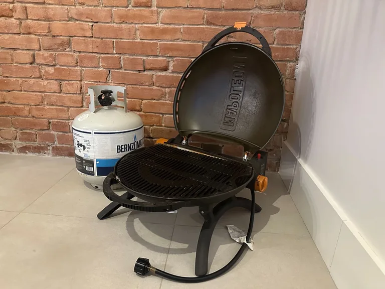 Barbecue portable NAPOLEON ouvert avec bonbonne de propane Bernzomatic, posé au sol devant un mur de briques