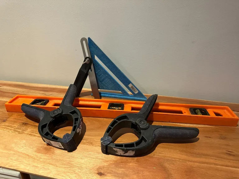 Kit d'outils de bricolage posé sur une table en bois : niveau orange, équerre bleue, fausse équerre métallique et deux serre-joints noirs