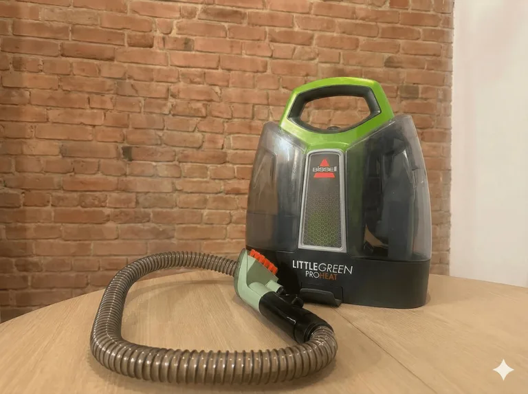 Nettoyeur portatif Bissell Little Green ProHeat posé sur une table en bois, avec un tuyau flexible enroulé, devant un mur de briques apparentes.