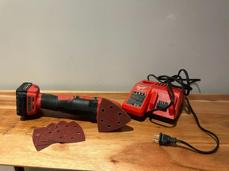 Ponceuse triangulaire sans fil Milwaukee avec chargeur et patins abrasifs grain 60, posée sur une table en bois